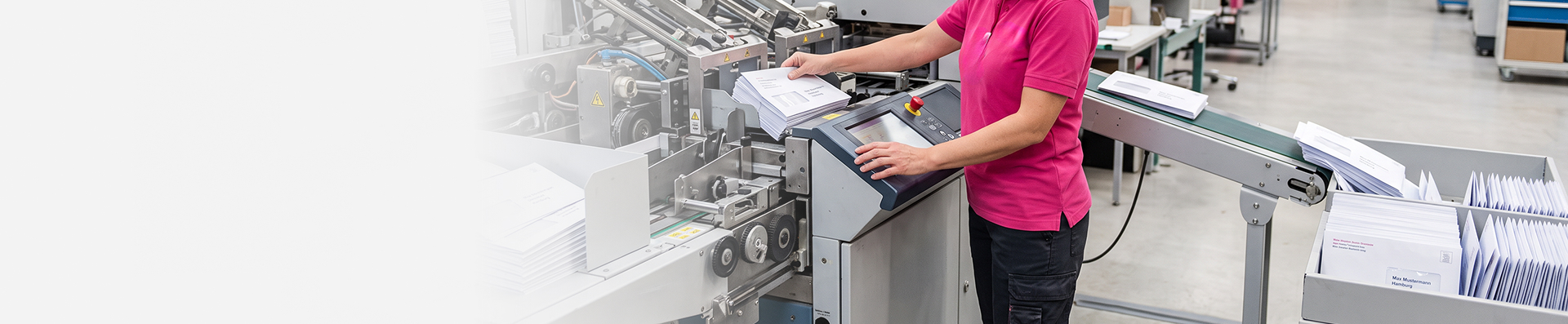Eine Mitarbeiterin bedient das Kontrollpanel einer Kuvertieranlage. Briefumschläge laufen über ein Förderband in Transportboxen für den Postversand.